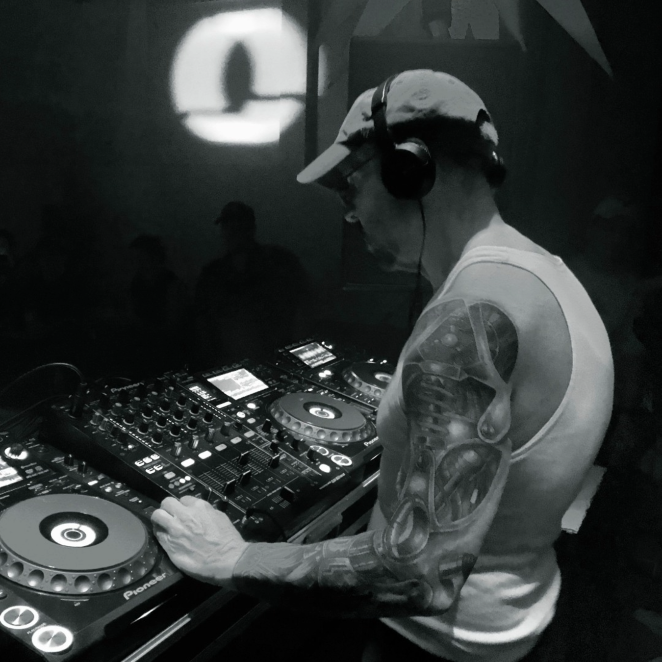 Black-and-white image of a DJ wearing headphones while mixing on turntables in a dimly lit booth.